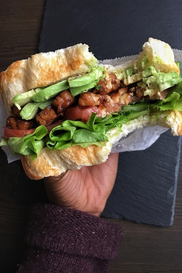 A hand holding a sandwich with avocado, tempeh and lettuce in it