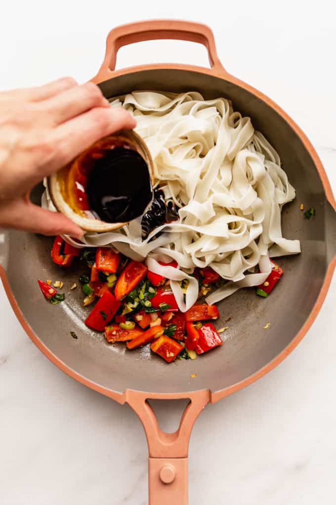 A hand pouring stir fry sauce into a pan of rice noodles