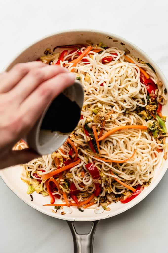 A hand pouring stir fry sauce into a pan of noodles and vegetables