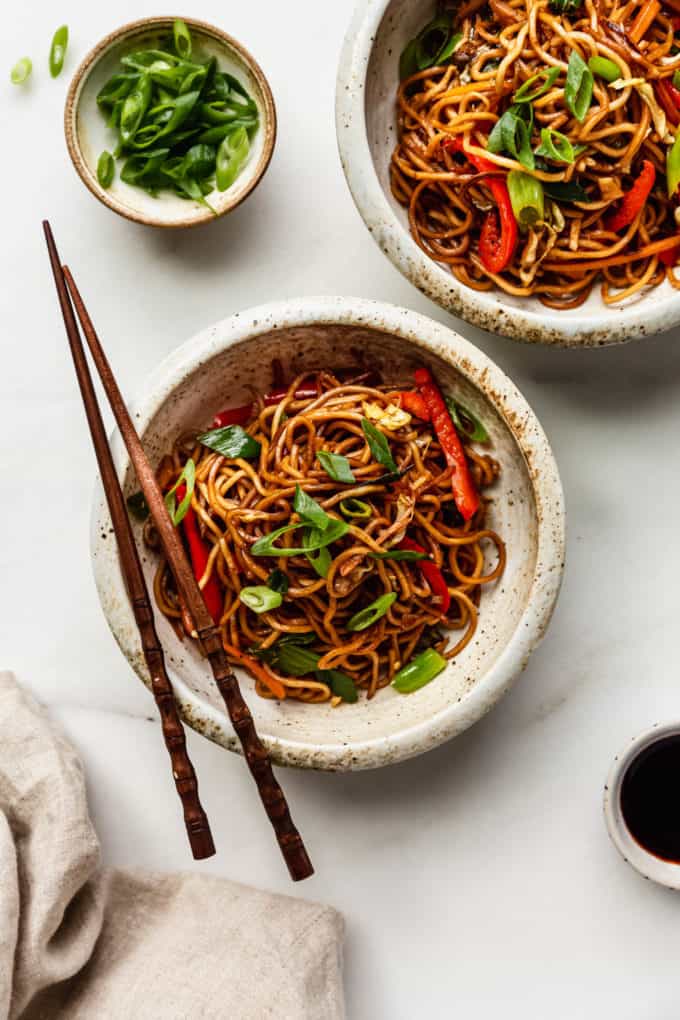 Two bowls of vegetable chow mein with a small bowl of green onions and a napkin on the side
