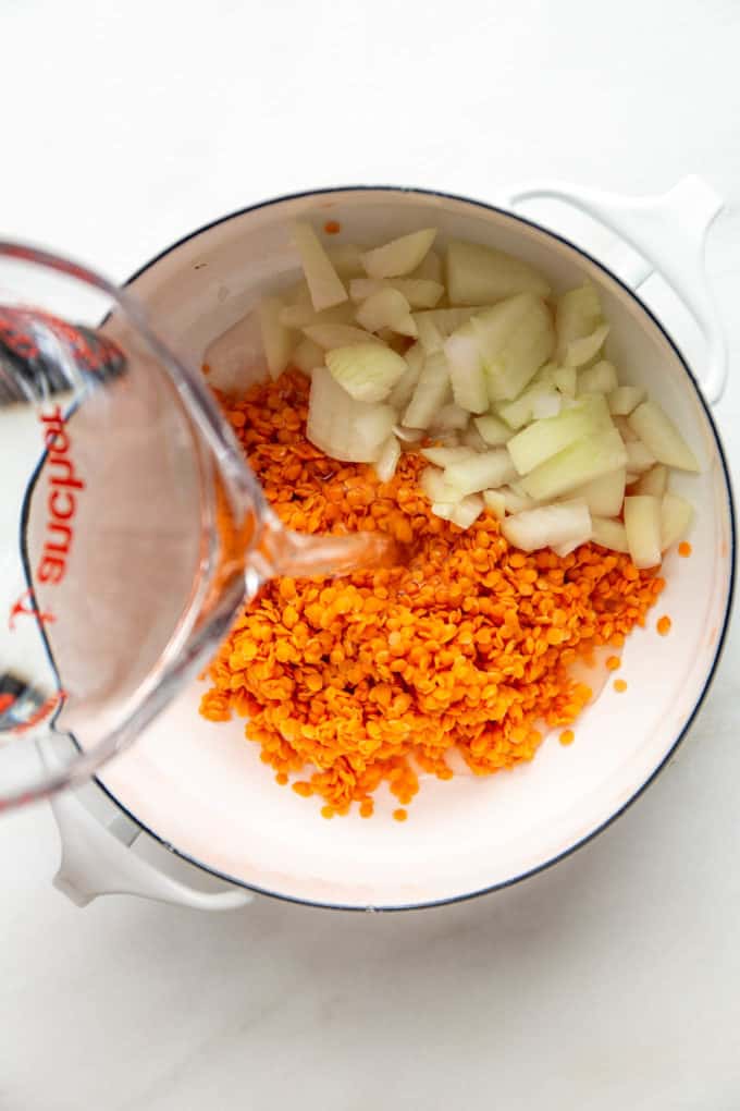 A measuring cup pouring water into a pot of red lentils and onions