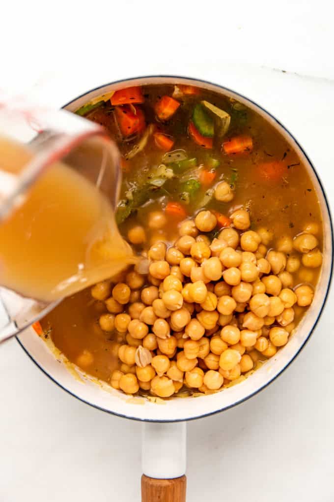 A measuring cup of broth being poured into a pot of chickpeas