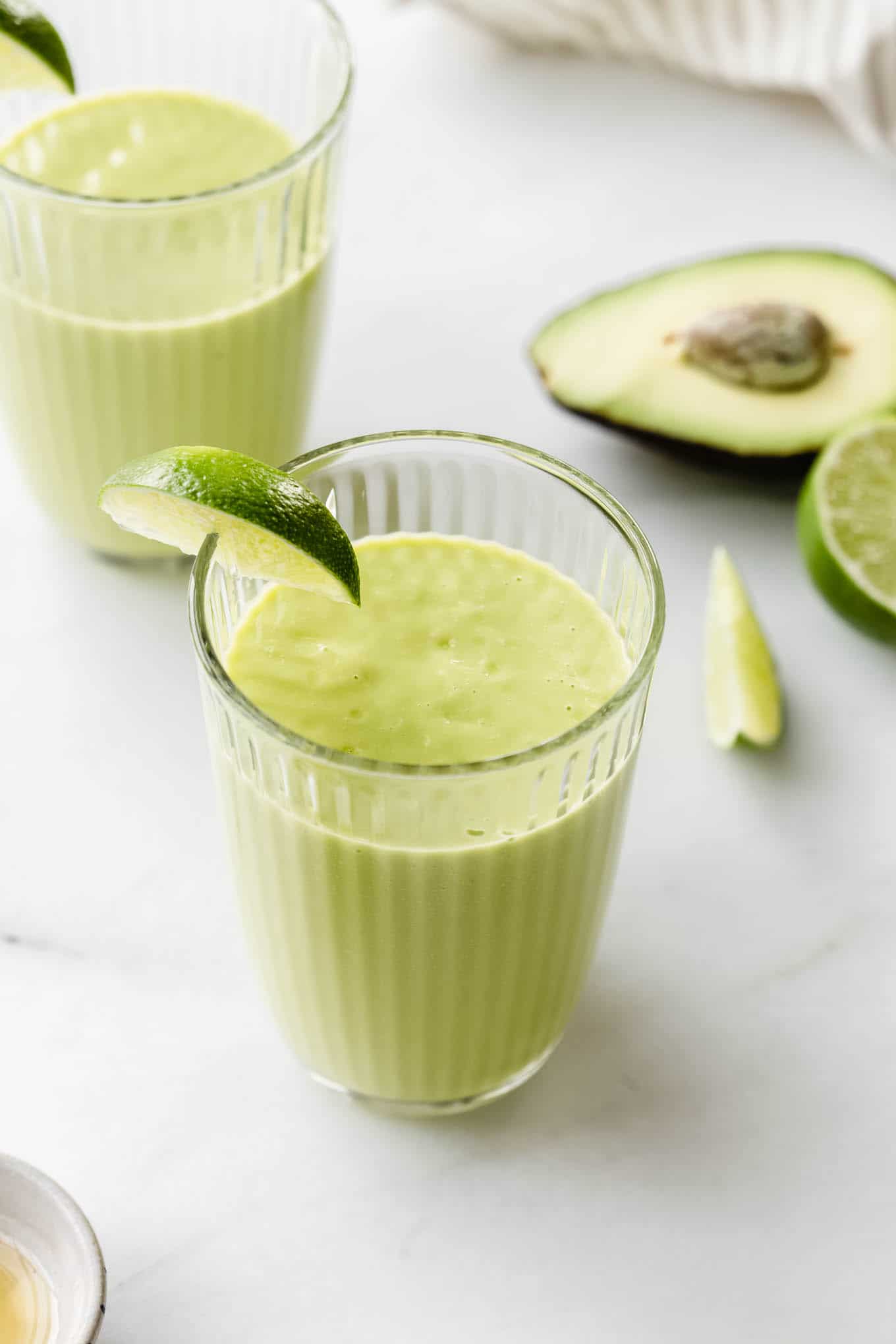 two avocado mango smoothies on a marble counter with sliced avocado and limes