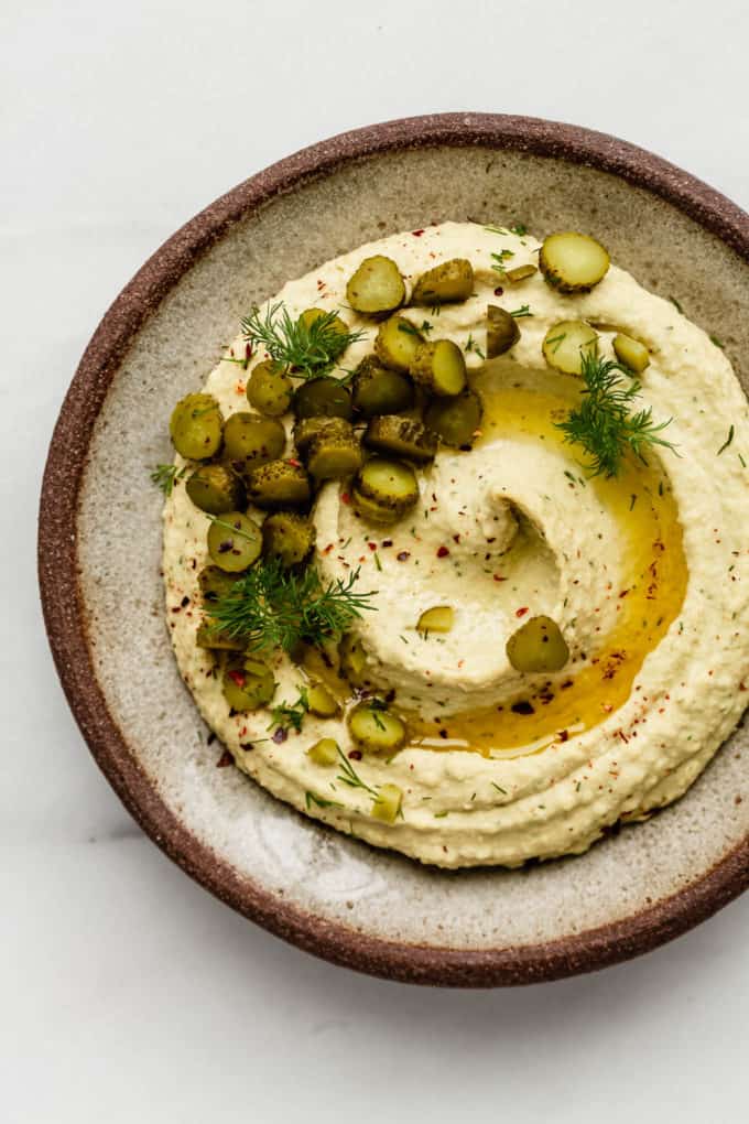 A ceramic plate with dill pickle hummus topped with pickles