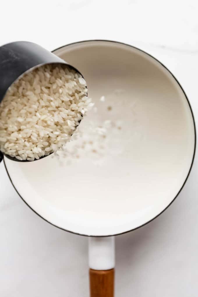 a measuring cup of rice being poured into a pot of coconut milk