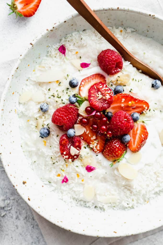 Coconut rice pudding topped with berries, help seeds and shredded coconut in a white speckled bowl