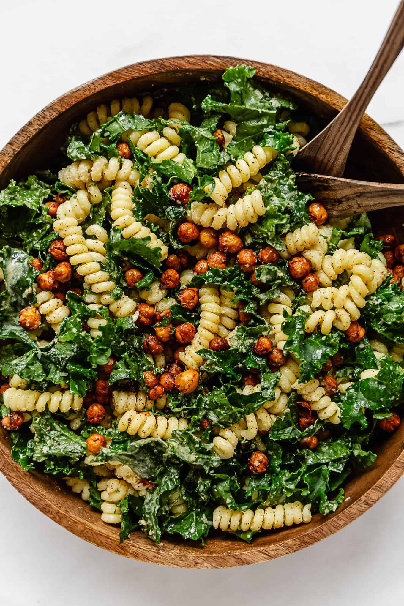 kale caesar pasta salad in a wood mixing bowl with 2 wood spoons