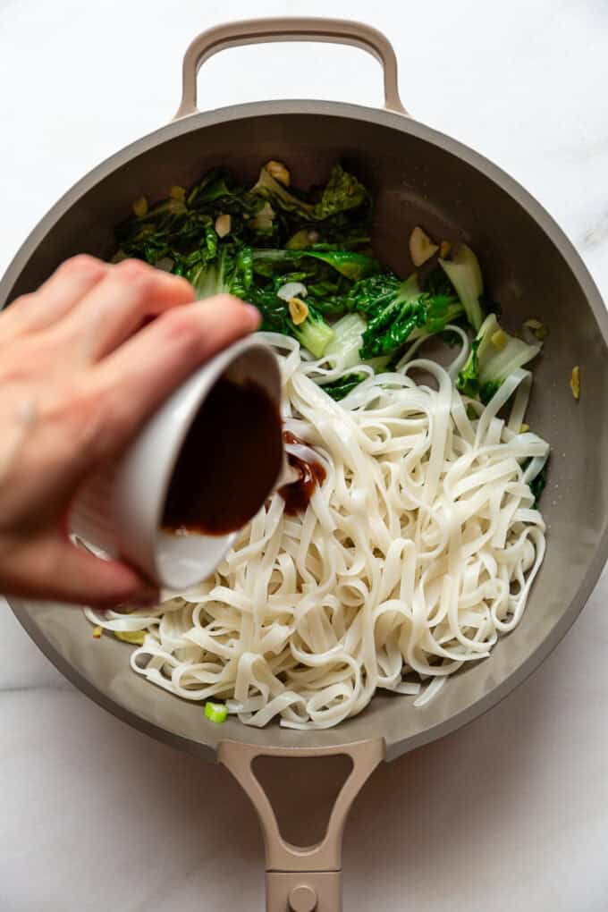 a hand pouring a cup of sauce into a pan of rice noodles and bok choy