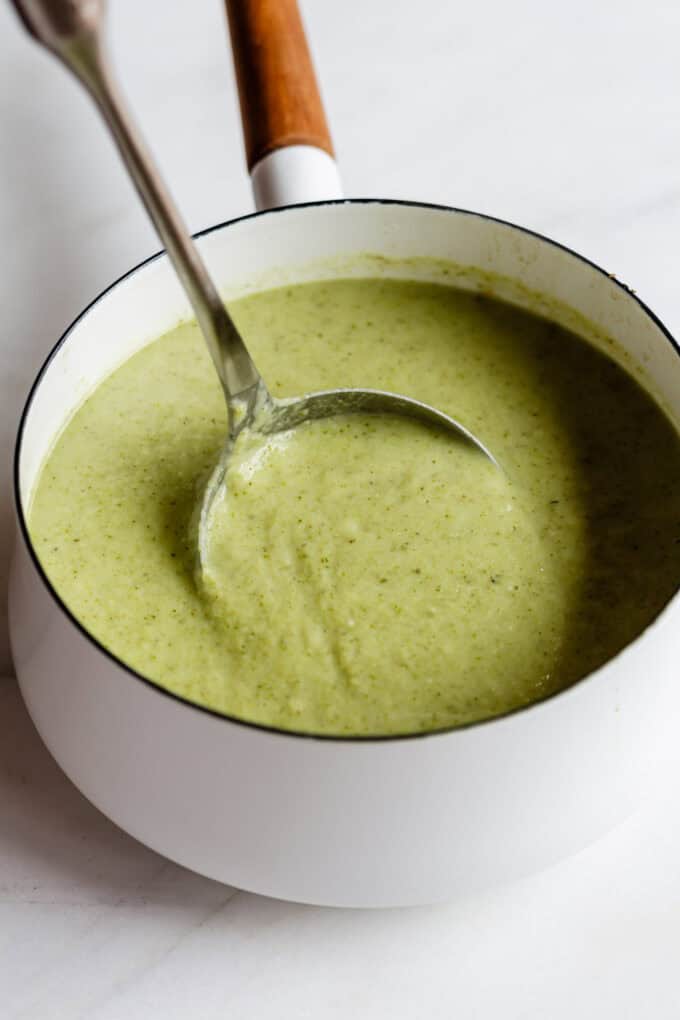 a ladle scooping broccoli cauliflower soup out of a white pot