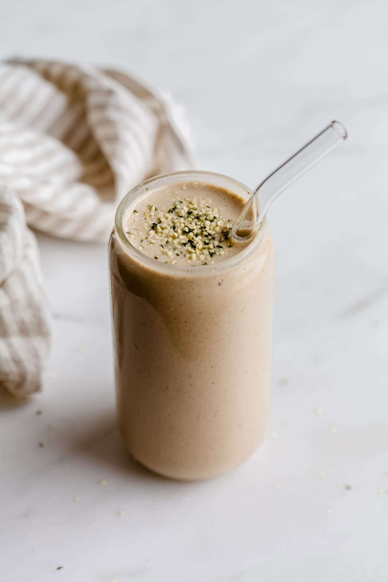 A banana date smoothie in a glass topped with hemp seeds and a striped beige napkin on the side.