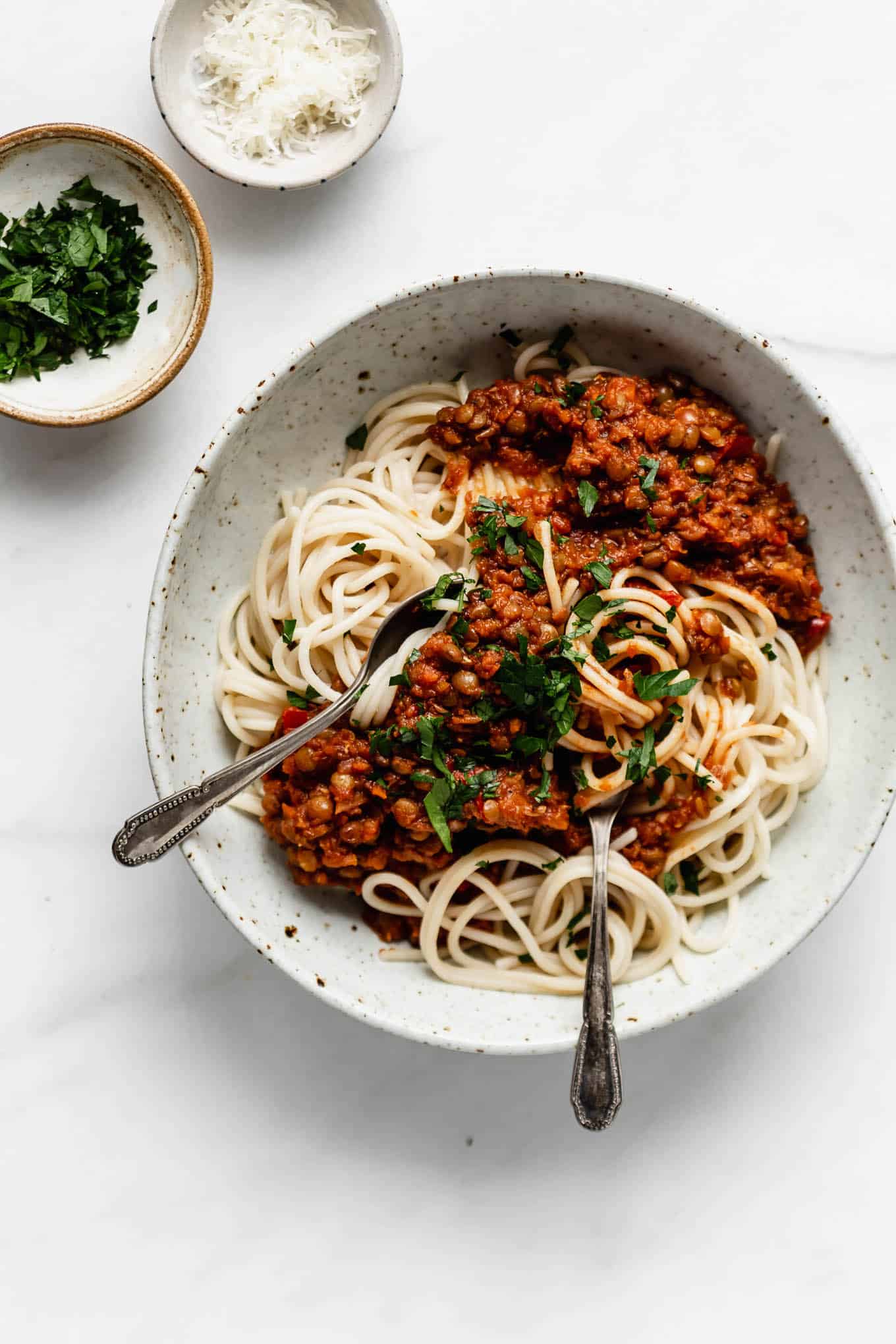 A bowl of spaghetti with lentil bolognese and parsley and parmesan on the side.