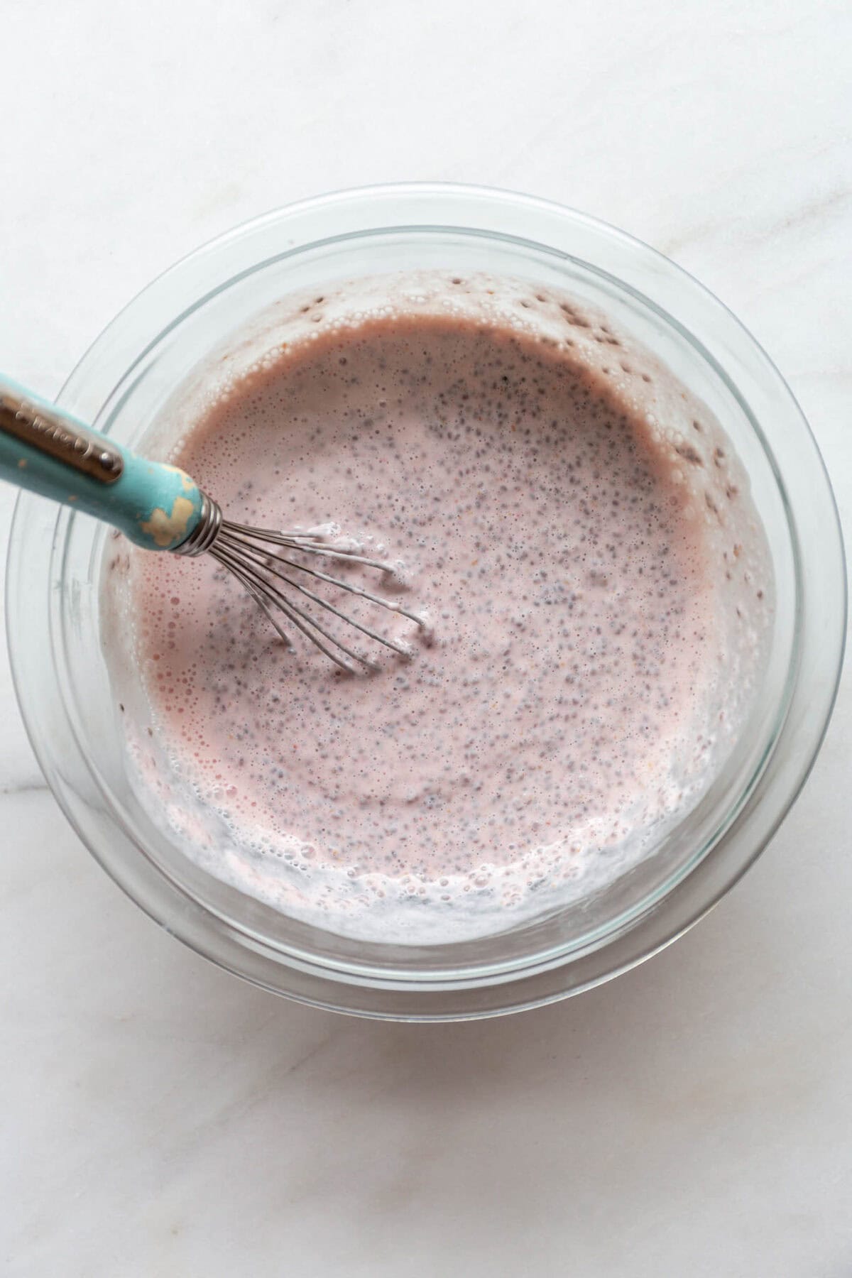 Pink chia pudding in a clear mixing bowl with a blue whisk.