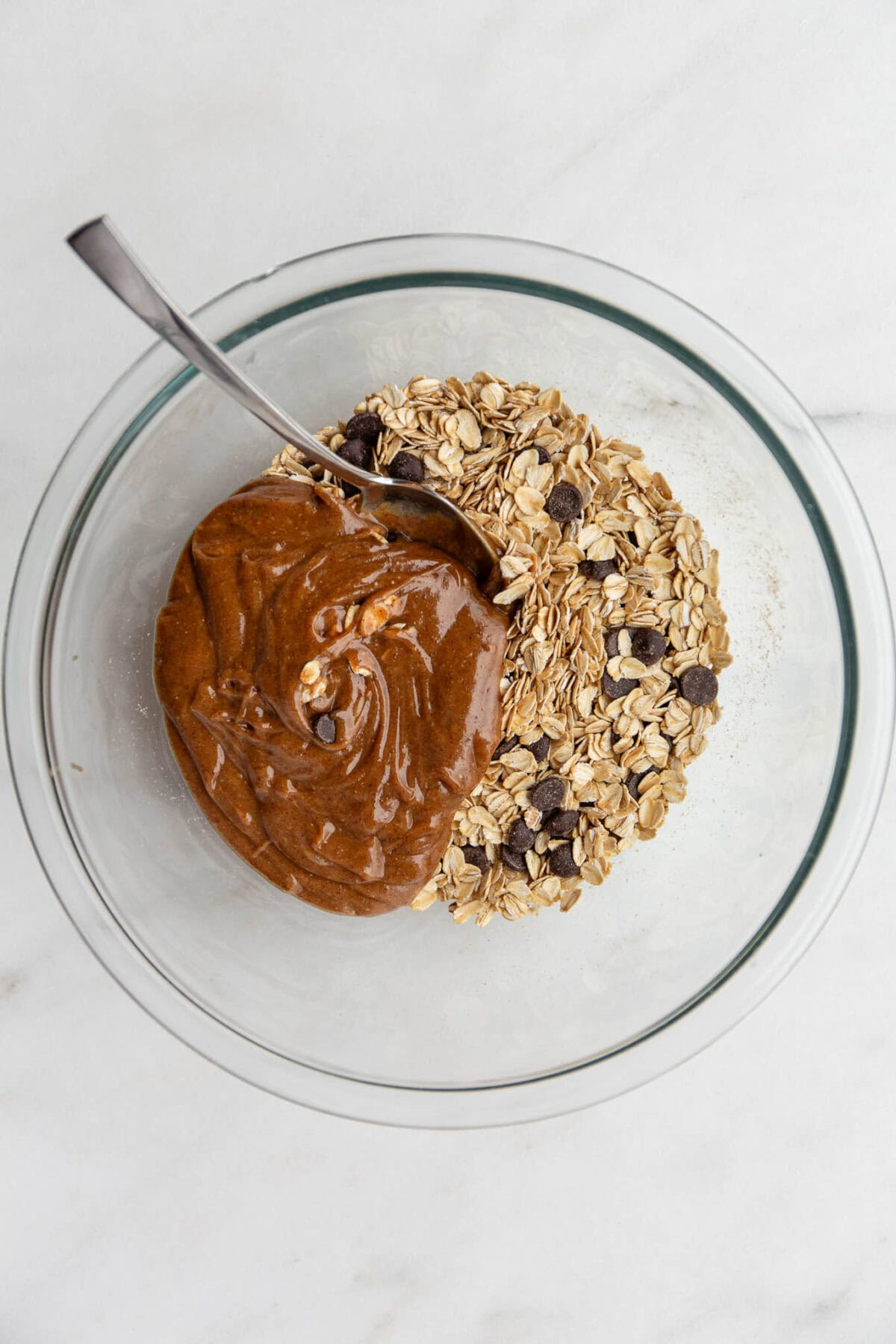 oats, chocolate chips, nut butter in a clear mixing bowl with a spoon.