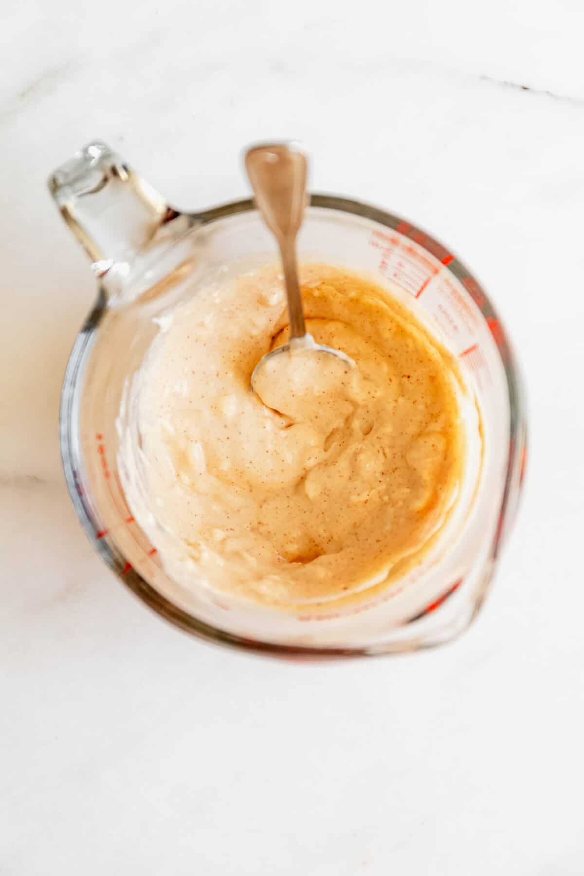 Cream cheese being stirred with a spoon in a glass measuring cup for the jalapeno popper chicken salad dressing.