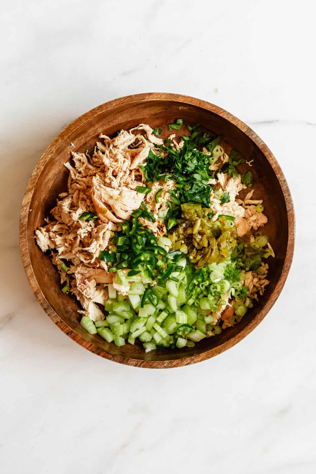Overhead view of shredded chicken and chopped celery and green onions in a wooden bowl for jalapeno popper chicken salad.