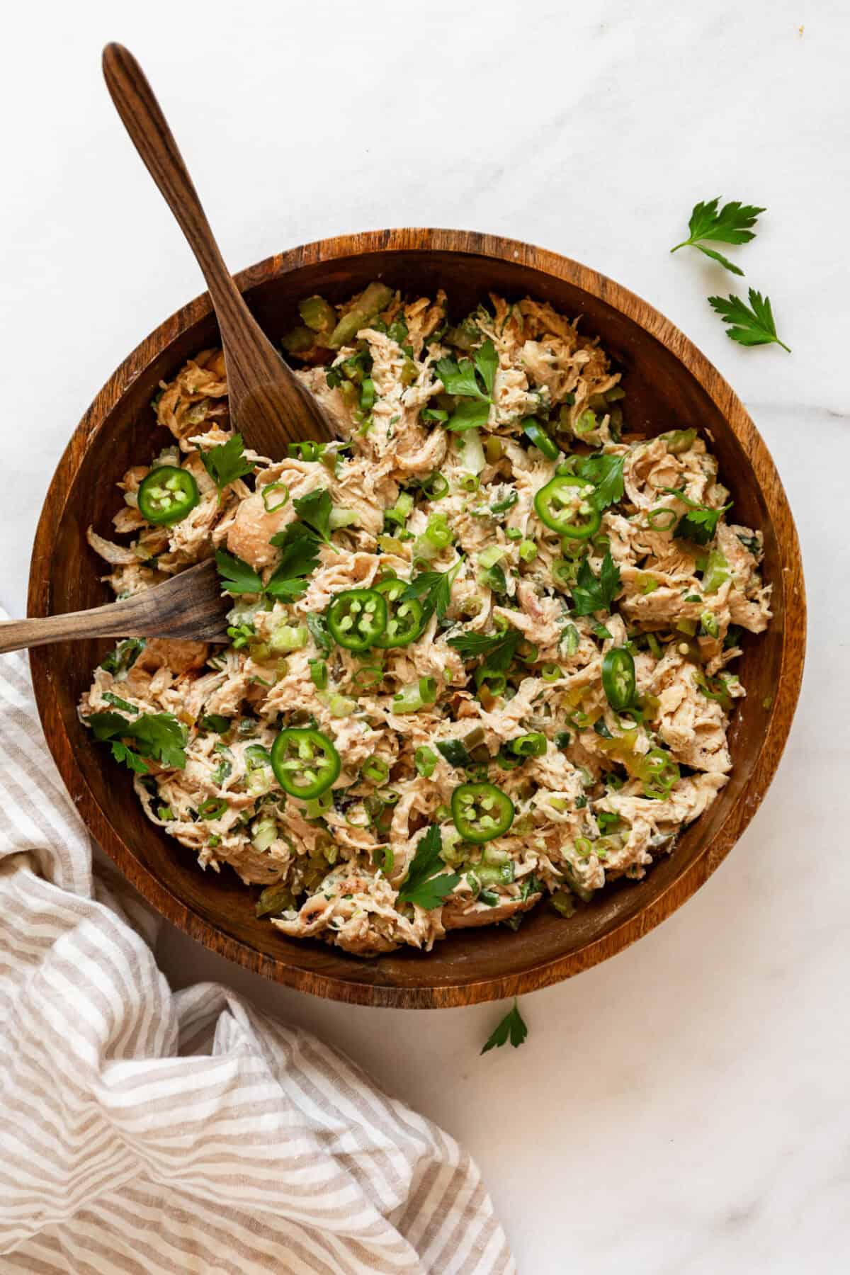 Overhead view of jalapeno popper chicken salad in a wooden bowl with a wooden spoon, topped with sliced jalapeños and fresh herbs.