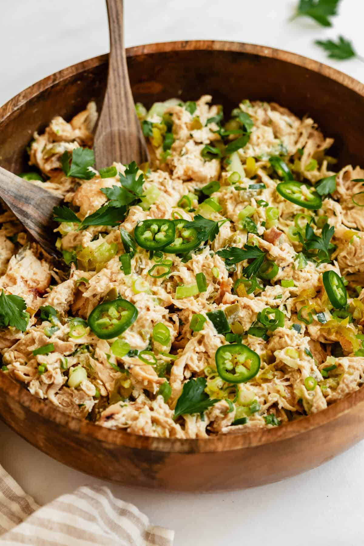 A close-up of jalapeno popper chicken salad in a wooden bowl, topped with sliced jalape&ntilde;os, green onions, and fresh herbs.