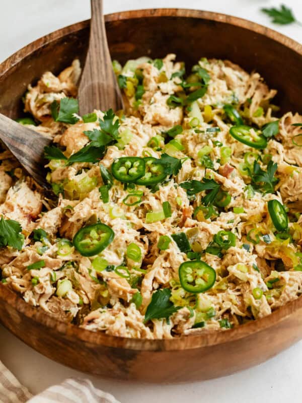 A close-up of jalapeno popper chicken salad in a wooden bowl, topped with sliced jalapeรฑos, green onions, and fresh herbs.