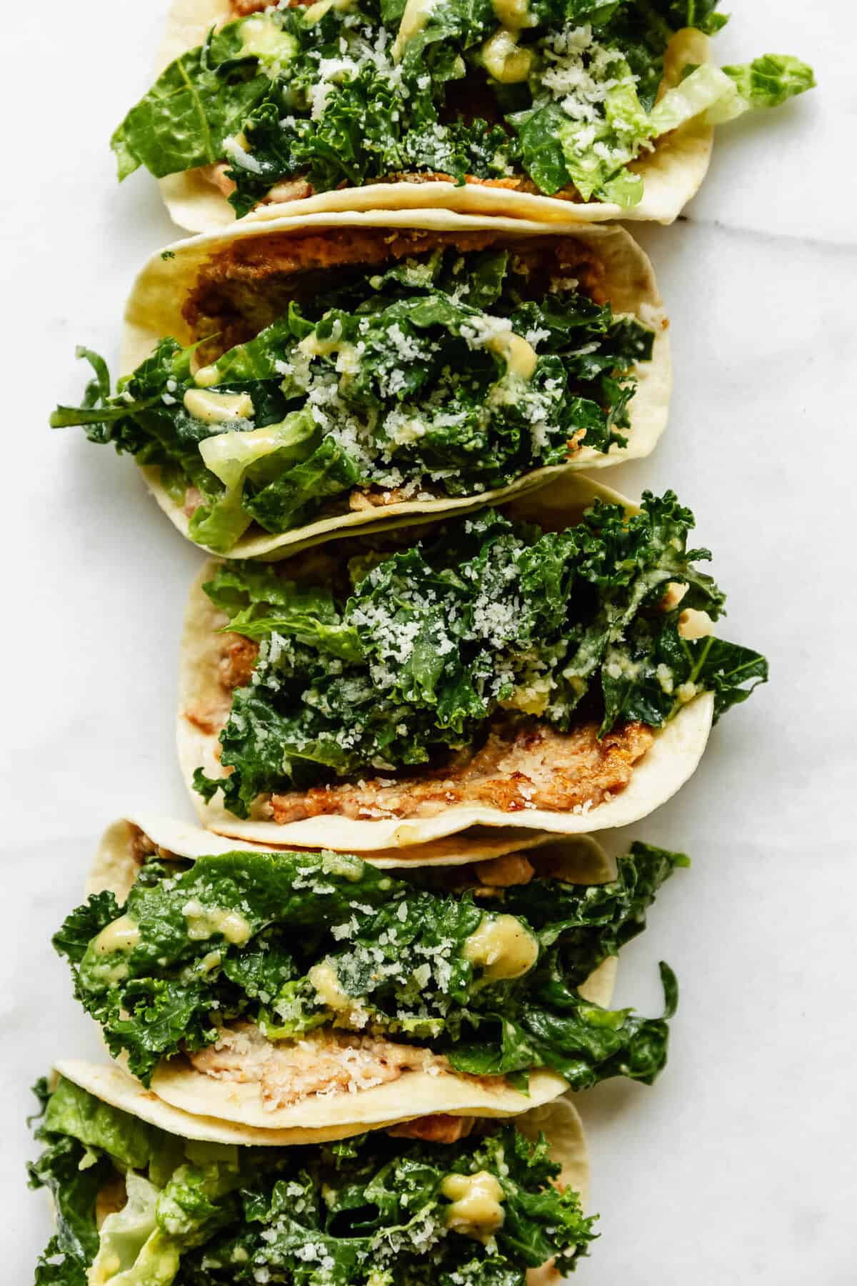 Three chicken caesar smashed tacos arranged in a row on white marble, topped with kale caesar salad and parmesan cheese.