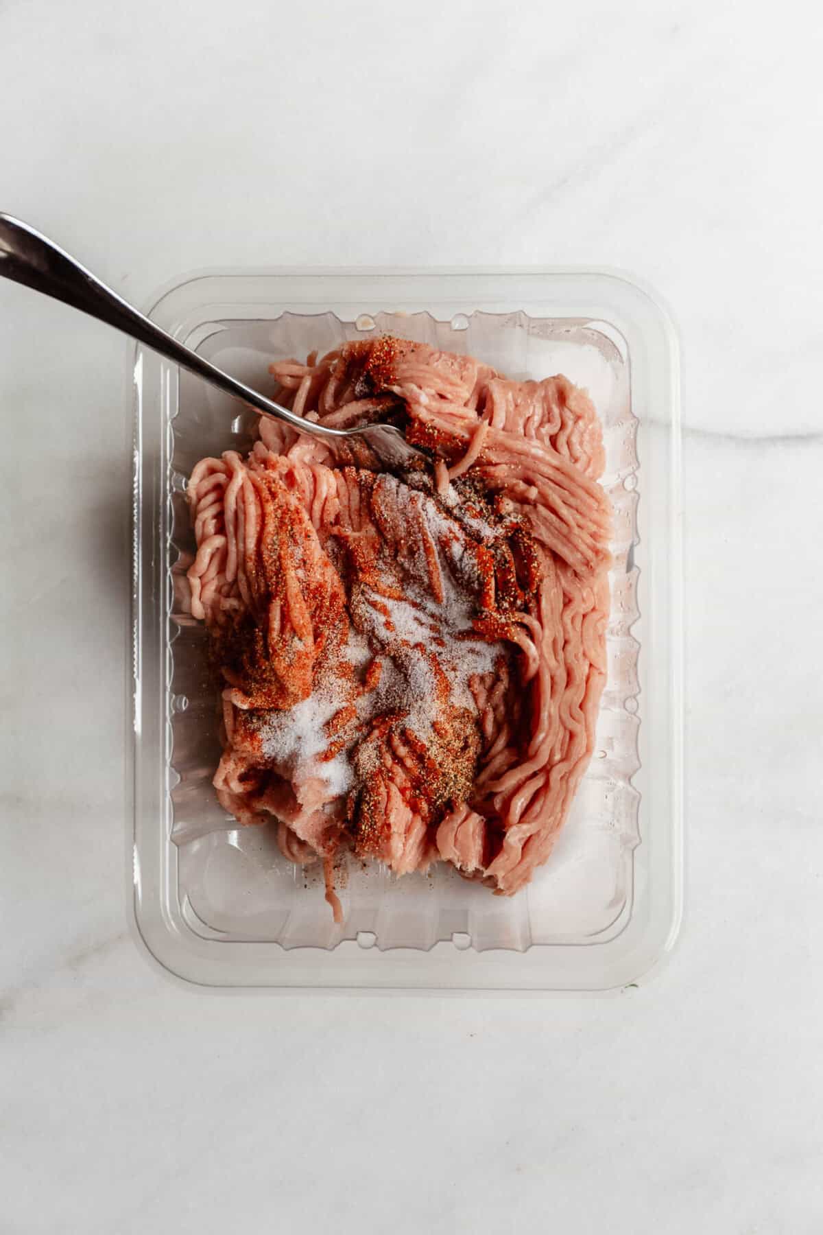 Raw ground chicken in a plastic container being seasoned with spices and mixed with a fork.