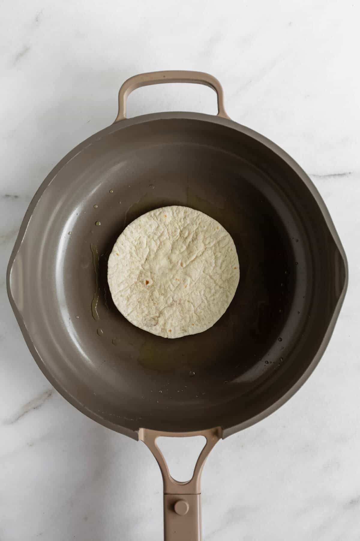 A flour tortilla in a dark skillet on a marble surface, ready to be toasted for chicken caesar smashed tacos.