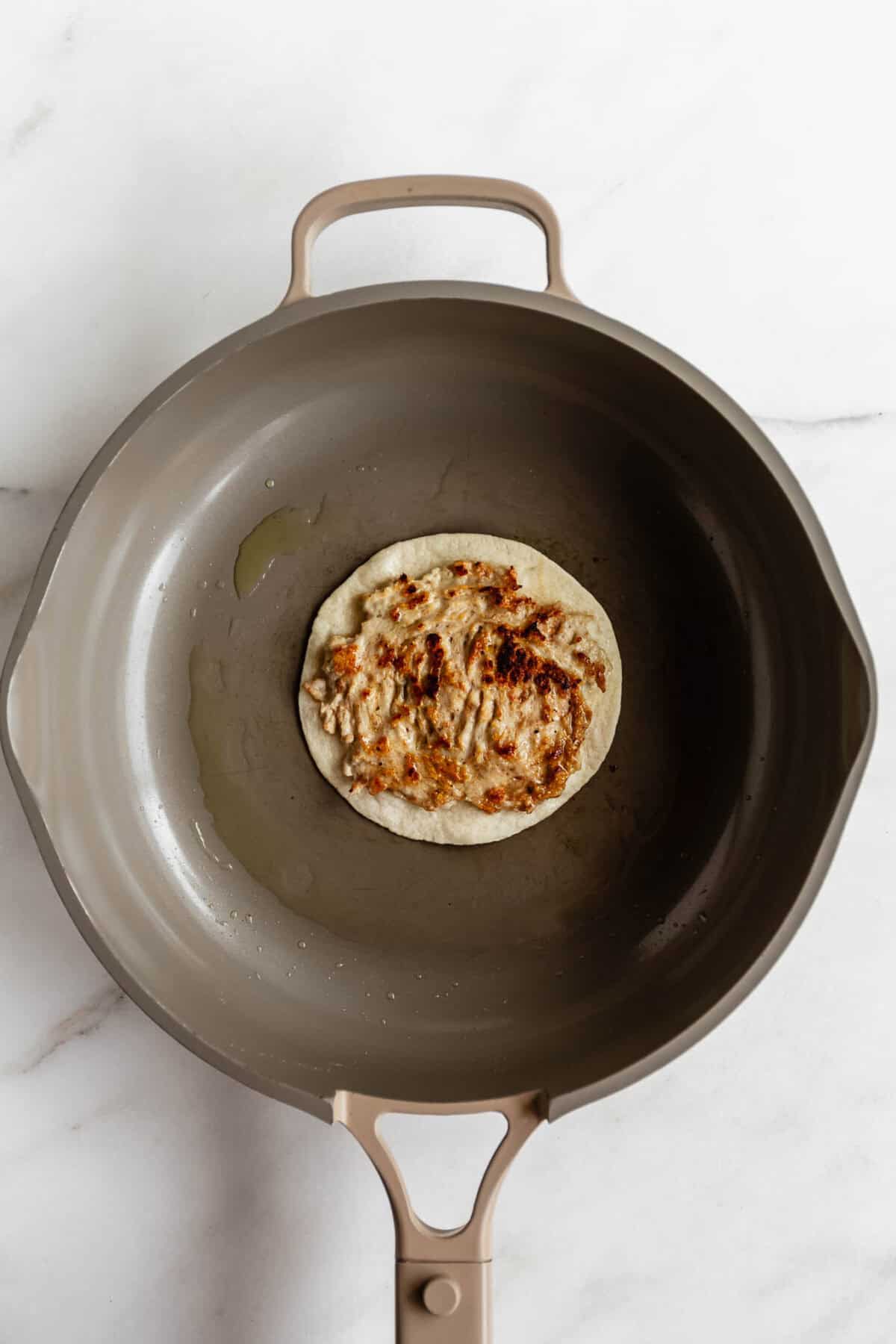 A toasted smashed tortilla in a dark skillet viewed from above, showing a golden brown crispy base for chicken caesar smashed tacos.