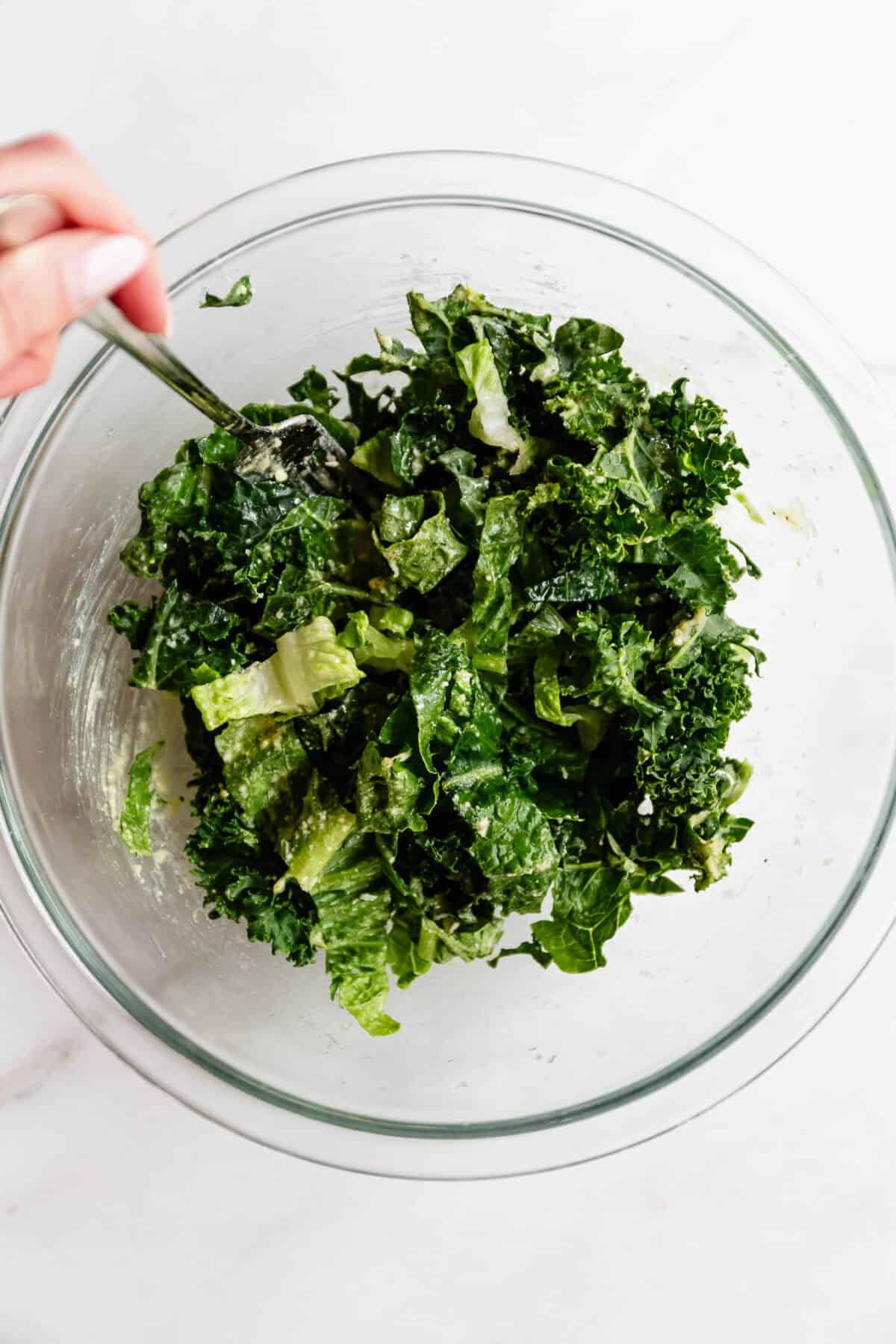 Hand tossing kale caesar salad with tongs in a glass bowl to prepare the topping for chicken caesar smashed tacos.