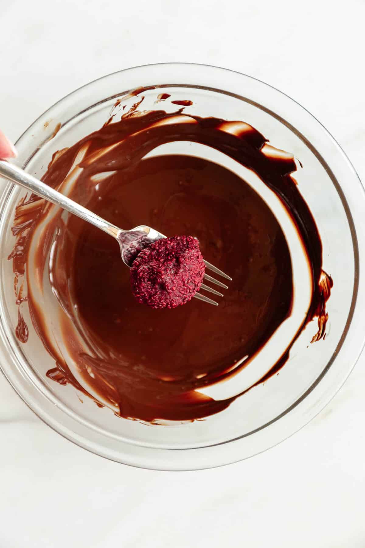a frozen raspberry chia jam bite on a fork above a bowl of dark chocolate.