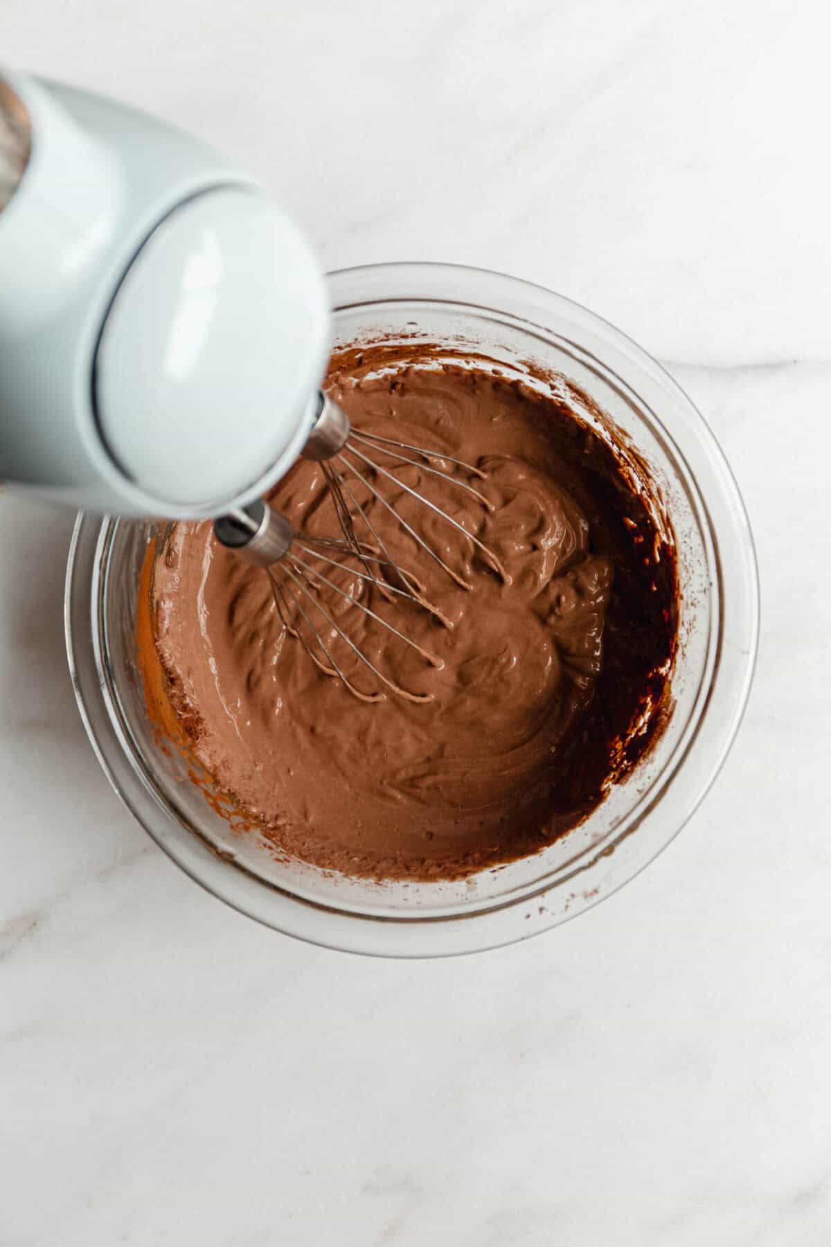 Using a hand mixer to blend Greek yogurt chocolate mousse ingredients in a glass bowl.