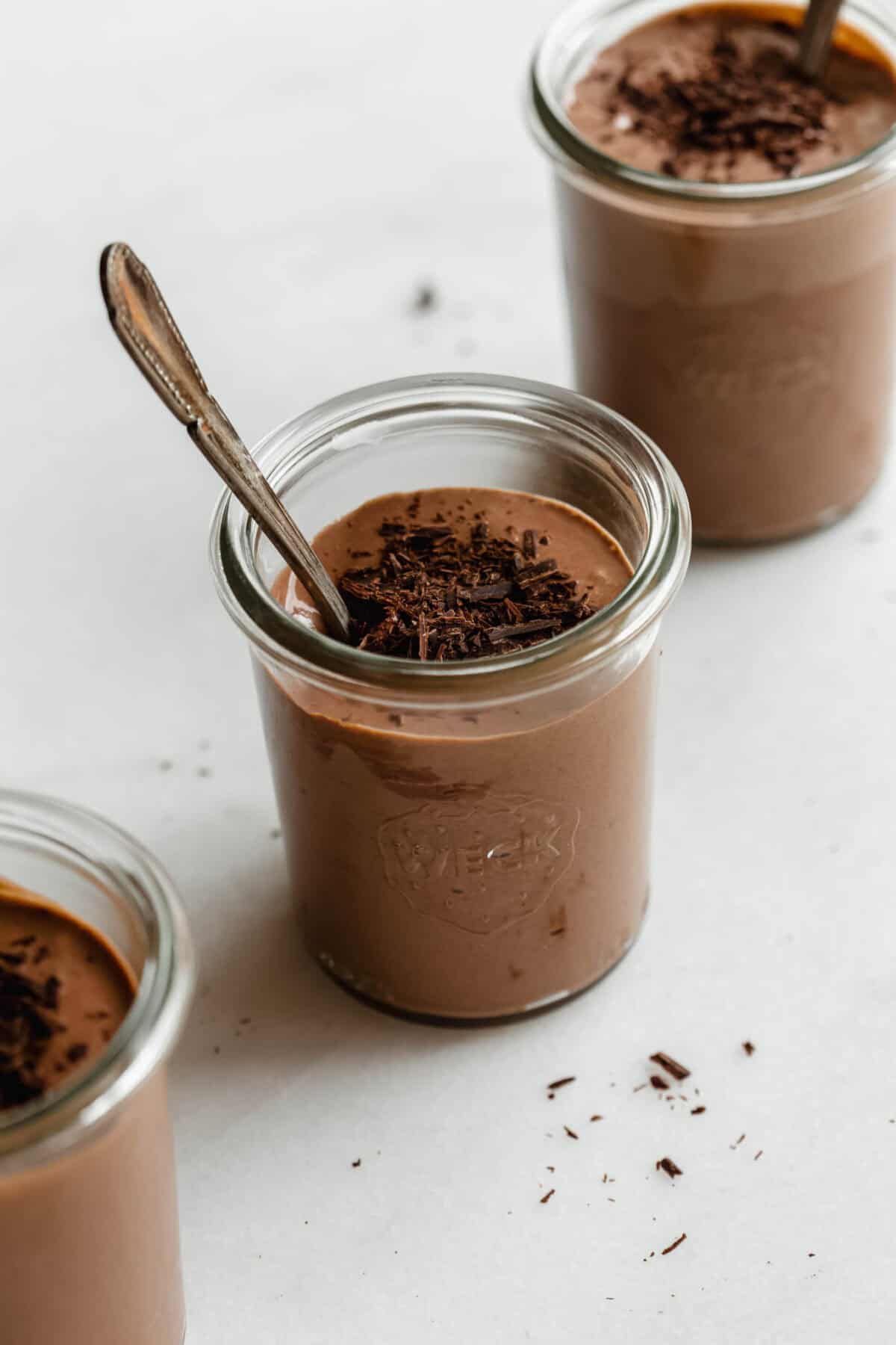 Multiple glass jars of Greek yogurt chocolate mousse topped with chocolate shavings on a white marble surface.