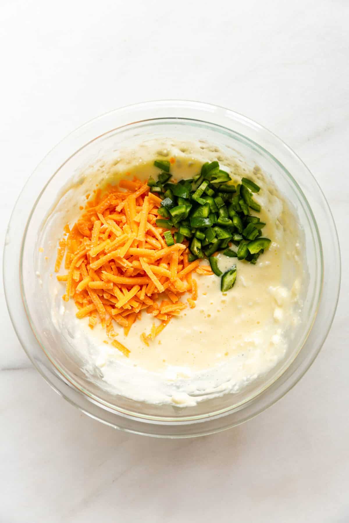 A bowl with shredded cheddar cheese and diced jalapeños being added to the cottage cheese biscuit mixture.