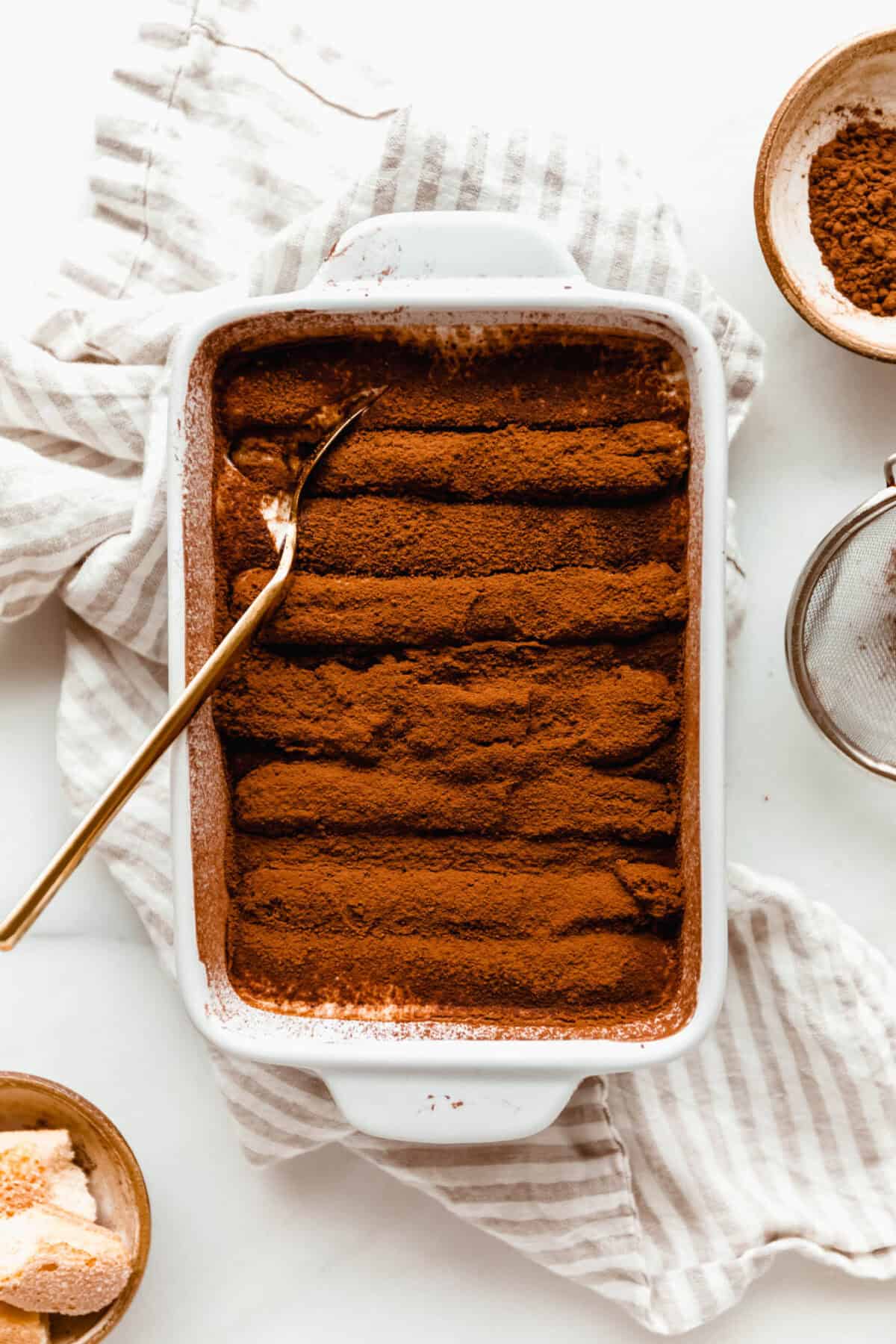 A finished Greek yogurt tiramisu in a white baking dish dusted with cocoa powder, served on a striped cloth.