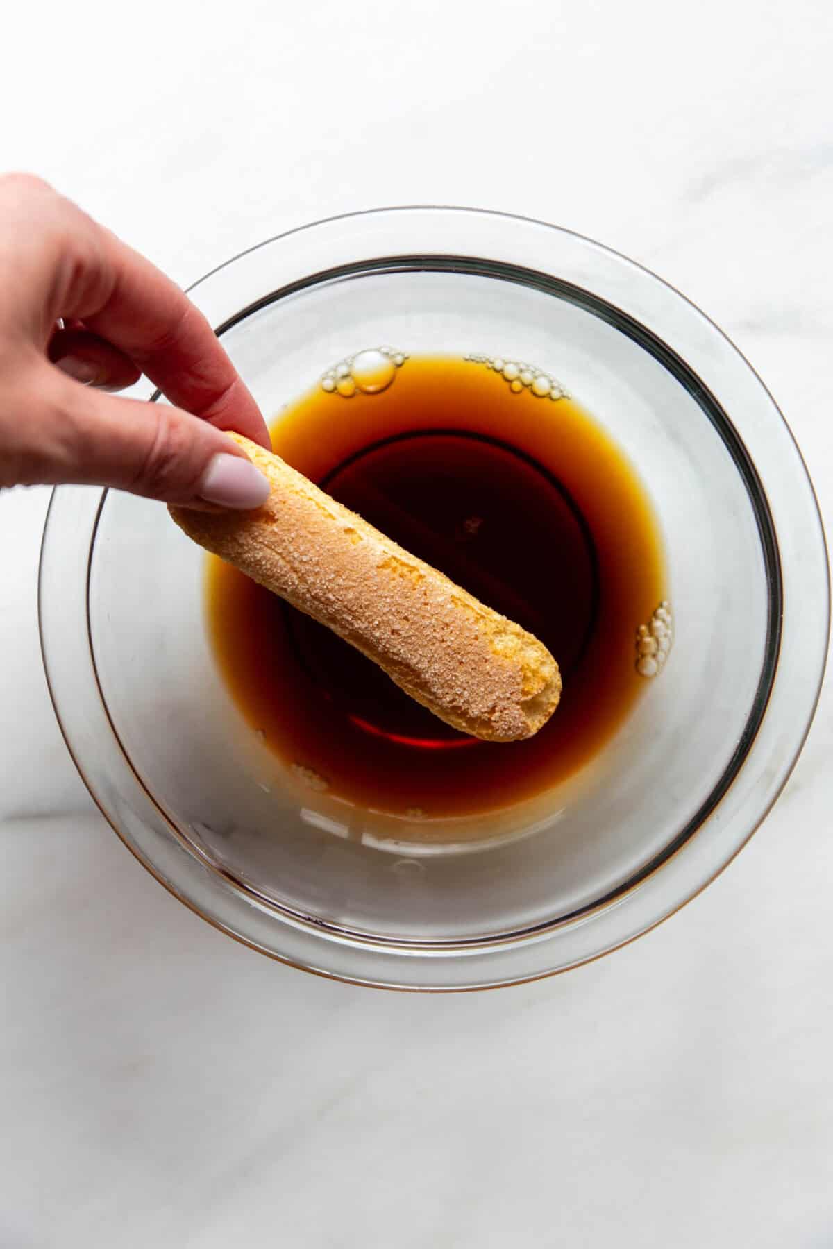 A hand dipping a ladyfinger biscuit into a bowl of espresso for Greek yogurt tiramisu.
