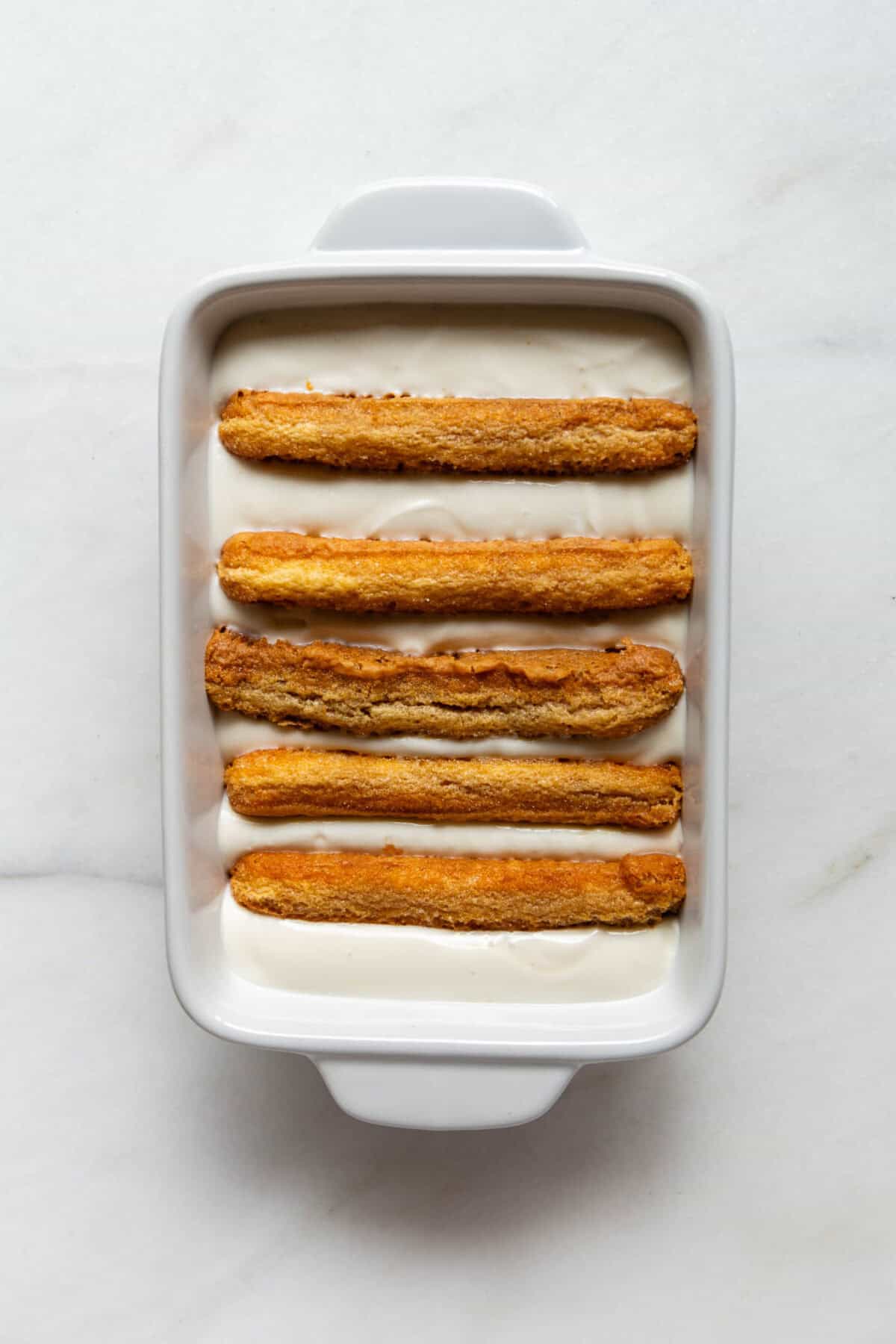 A white baking dish with a layer of espresso-soaked ladyfinger biscuits and Greek yogurt filling.