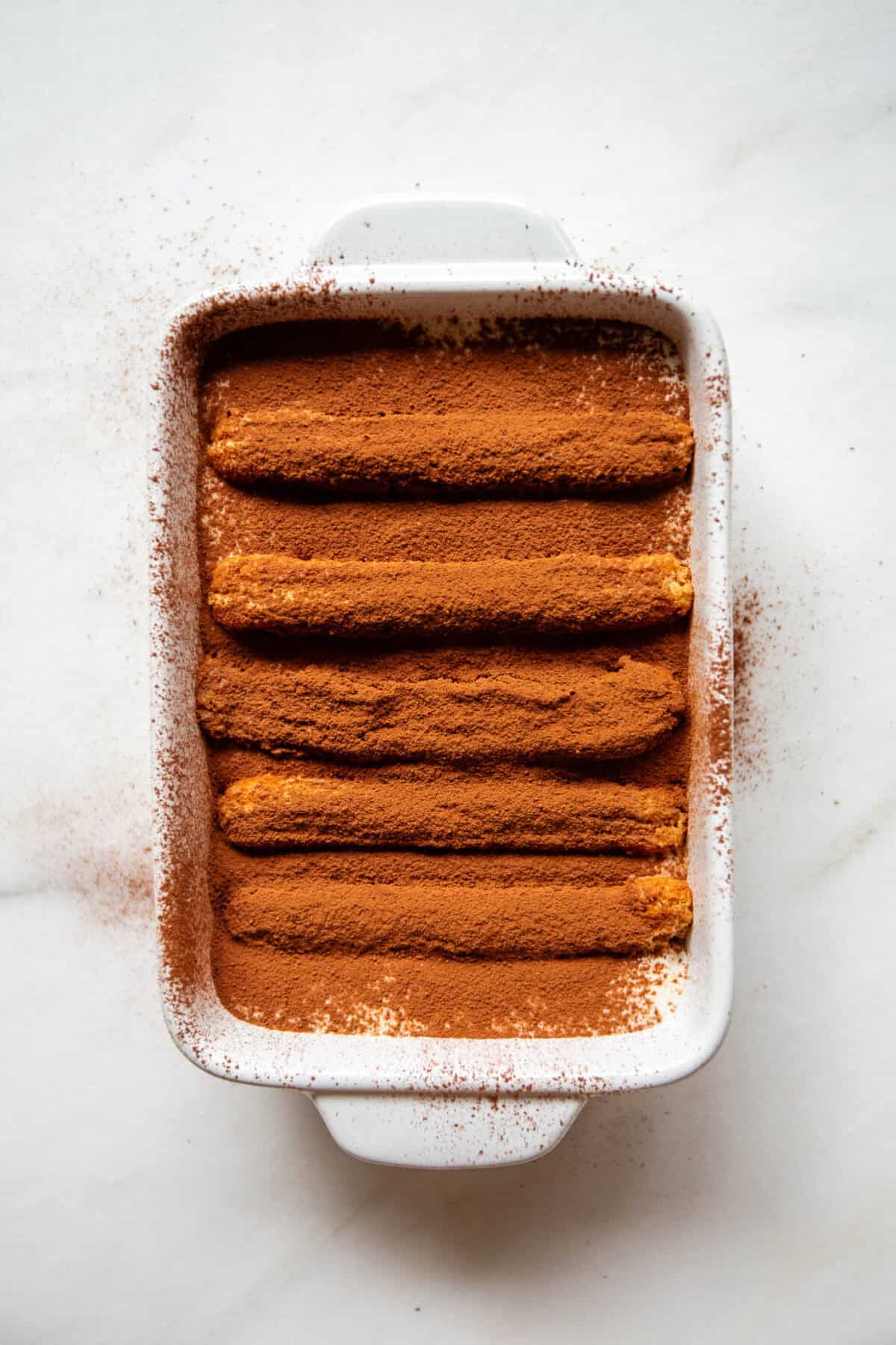 A white baking dish filled with Greek yogurt tiramisu generously dusted with cocoa powder