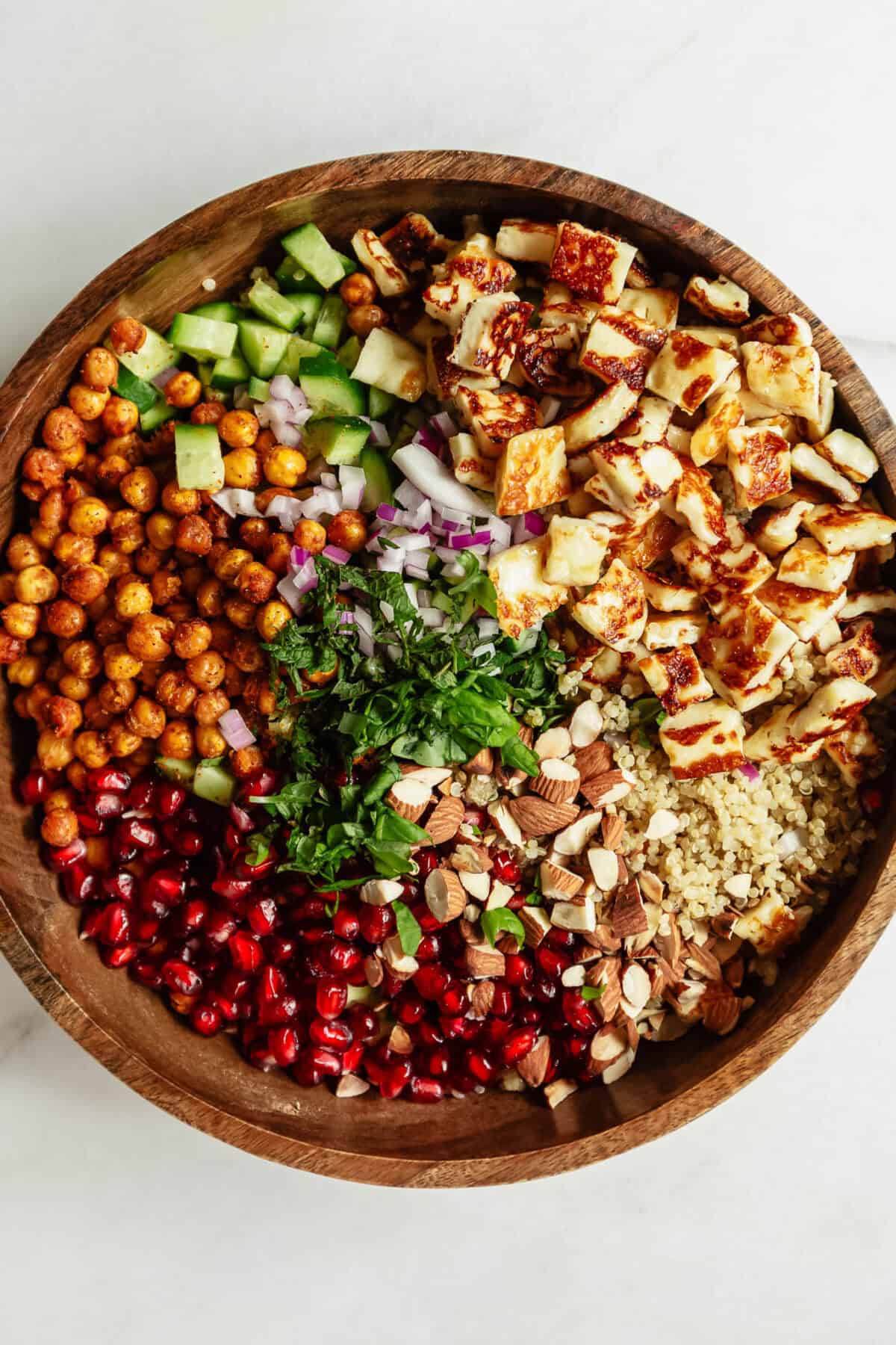 quinoa, roasted chickpeas, cucumber, halloumi, pomegranate, red onion and herbs in a wood salad bowl.