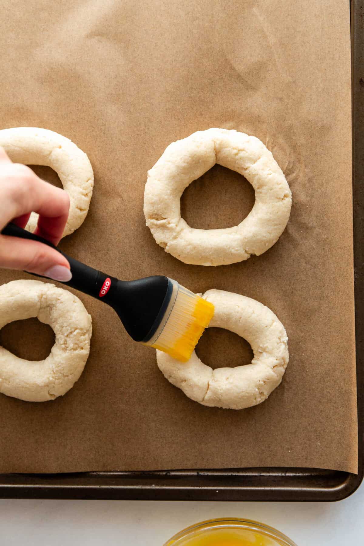 A hand brushing egg wash onto unbaked Cottage Cheese Almond Flour Bagels.