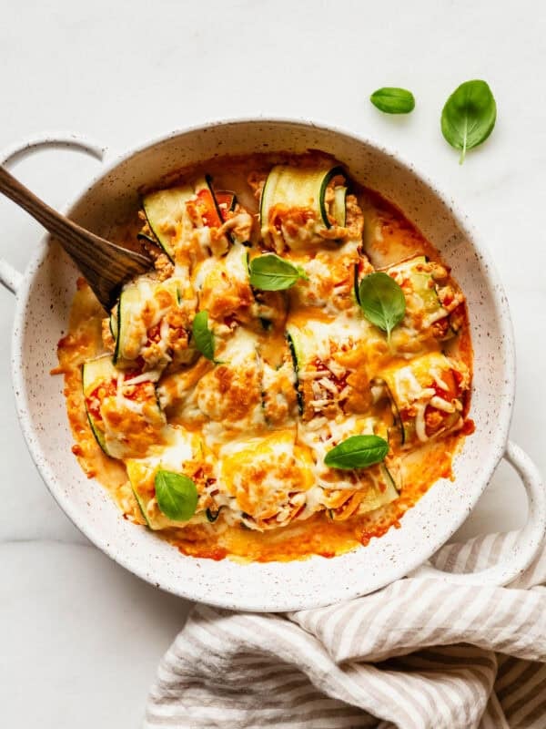 buffalo chicken zucchini roll ups in a baking dish with a beige stripe napkin on the side.