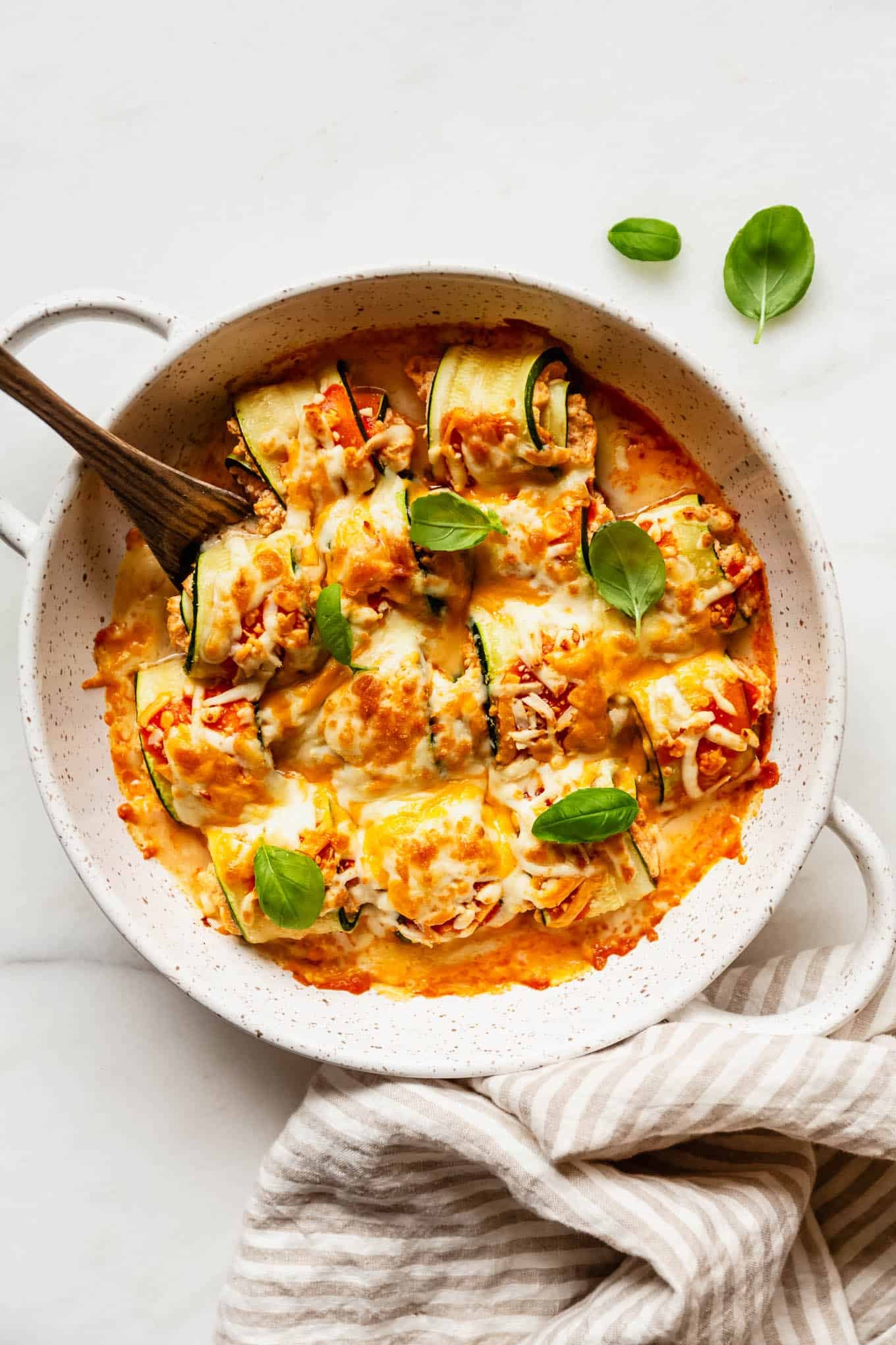 buffalo chicken zucchini roll ups in a baking dish with a beige stripe napkin on the side.