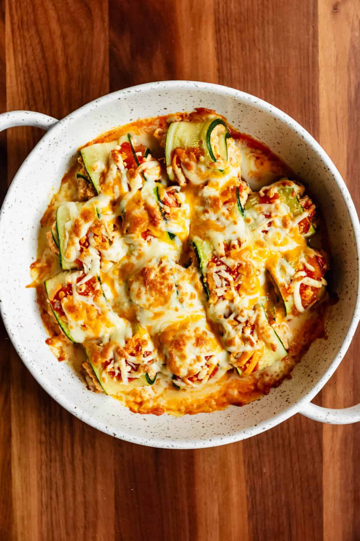 baked buffalo chicken zucchini roll ups in a baking dish with hot sauce topped with melted shredded cheeese.