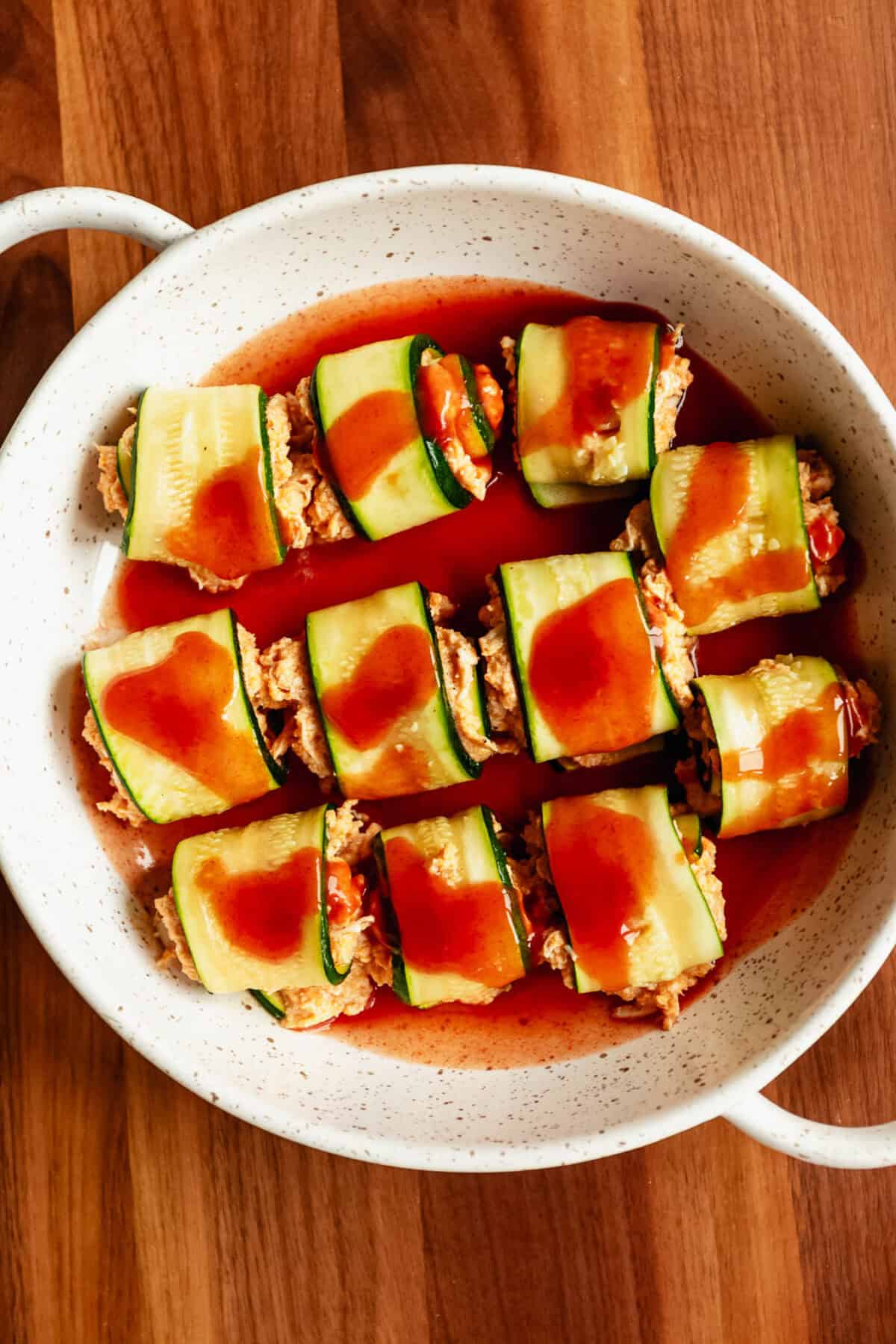 buffalo chicken zucchini roll ups in a baking dish with hot sauce.