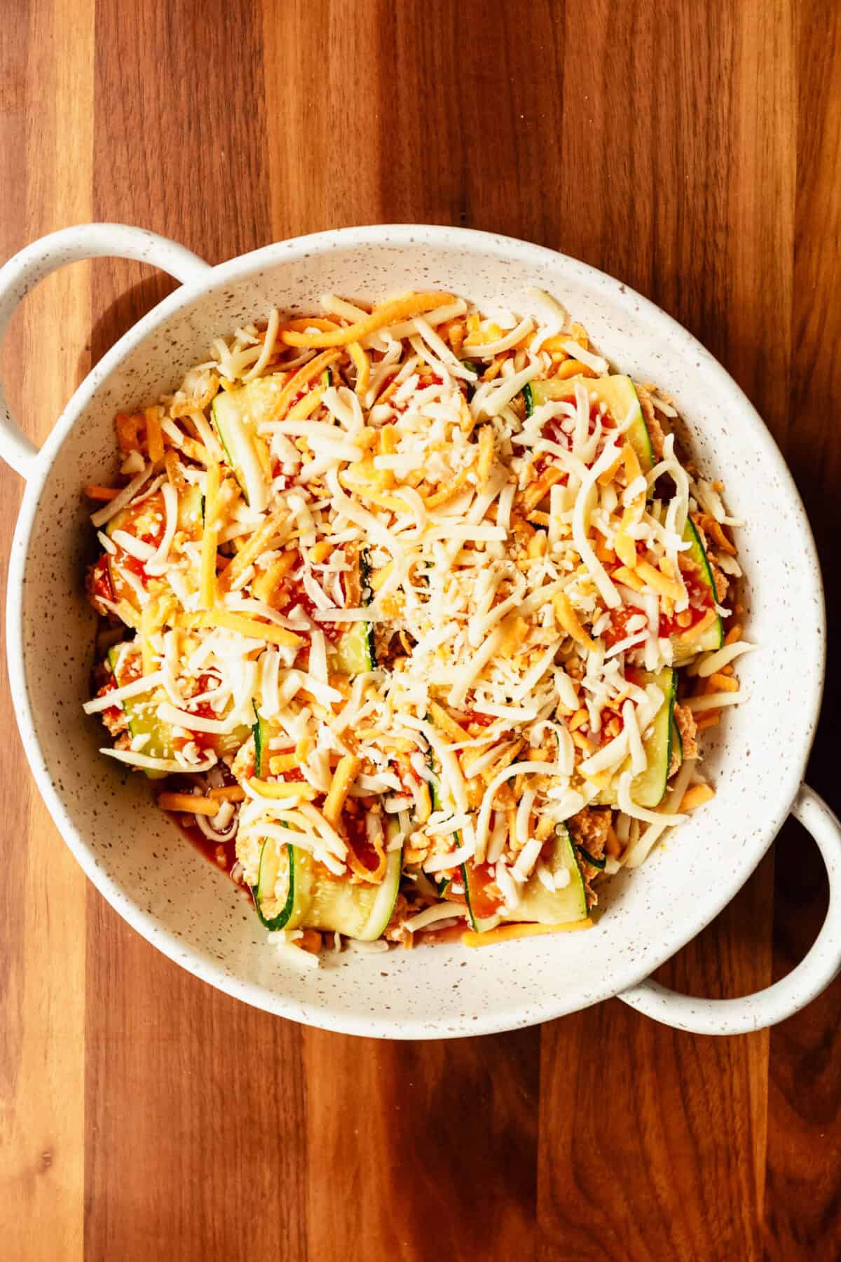 buffalo chicken zucchini roll ups in a baking dish with hot sauce topped with shredded cheese.