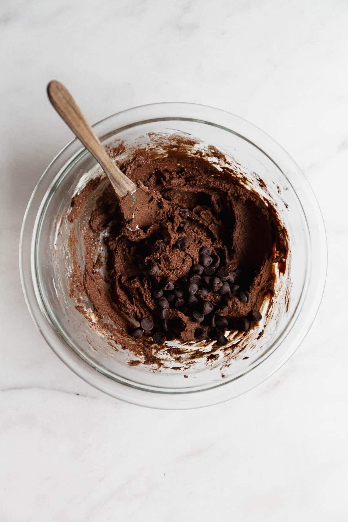 Cottage cheese brownie batter in a clear mixing bowl with a wood spoon and chocolate chips.