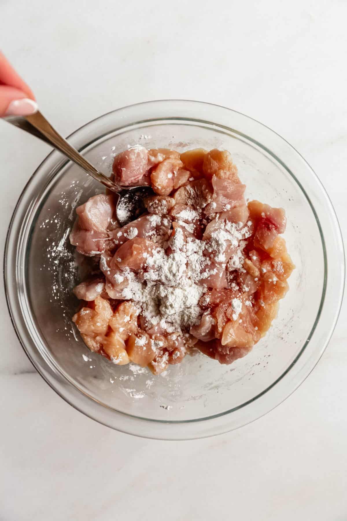 A hand stirring diced raw chicken with cornstarch in a glass mixing bowl.
