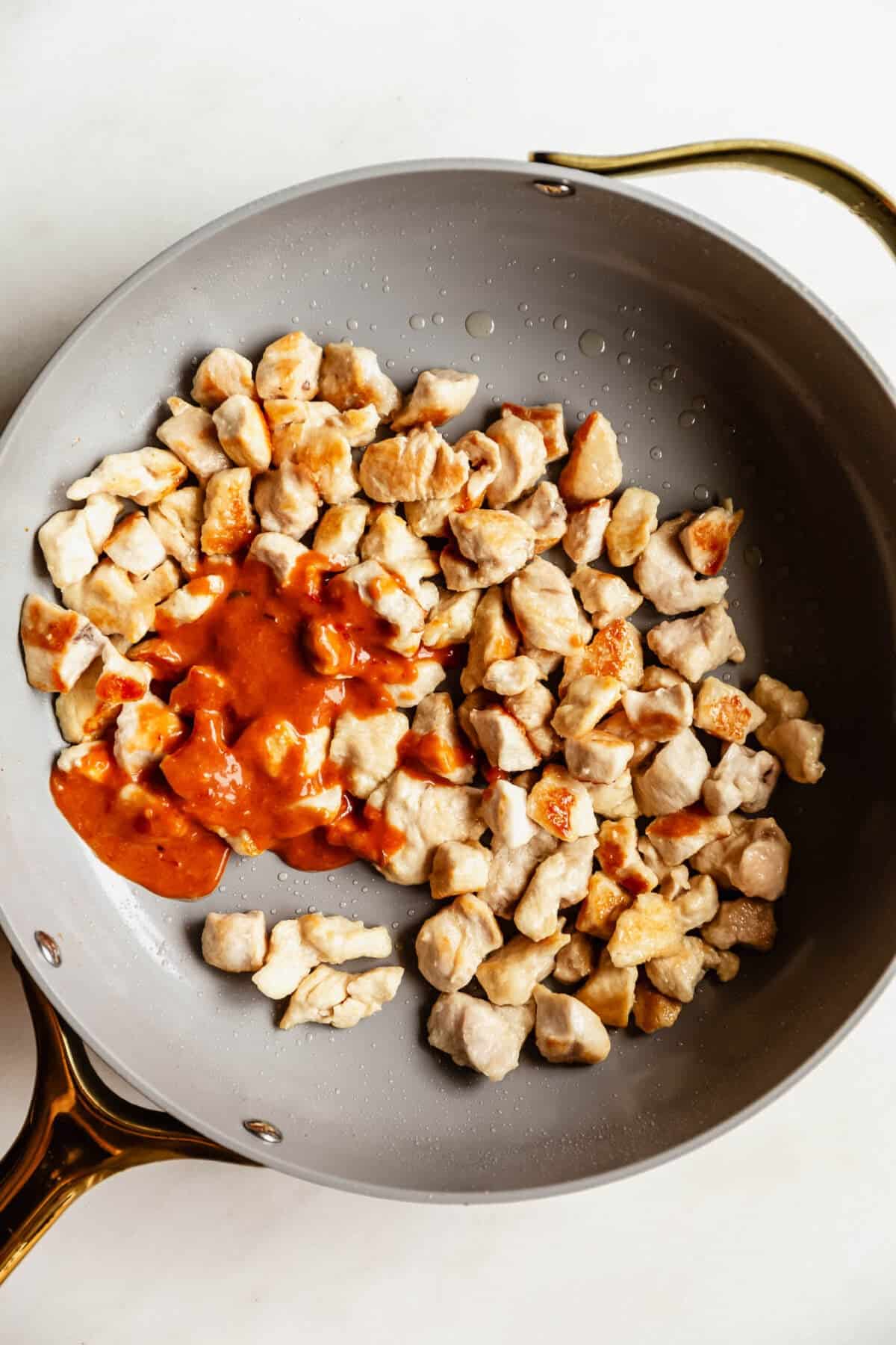Diced raw chicken pieces searing in a skillet with bang bang sauce being poured over half of the pan.