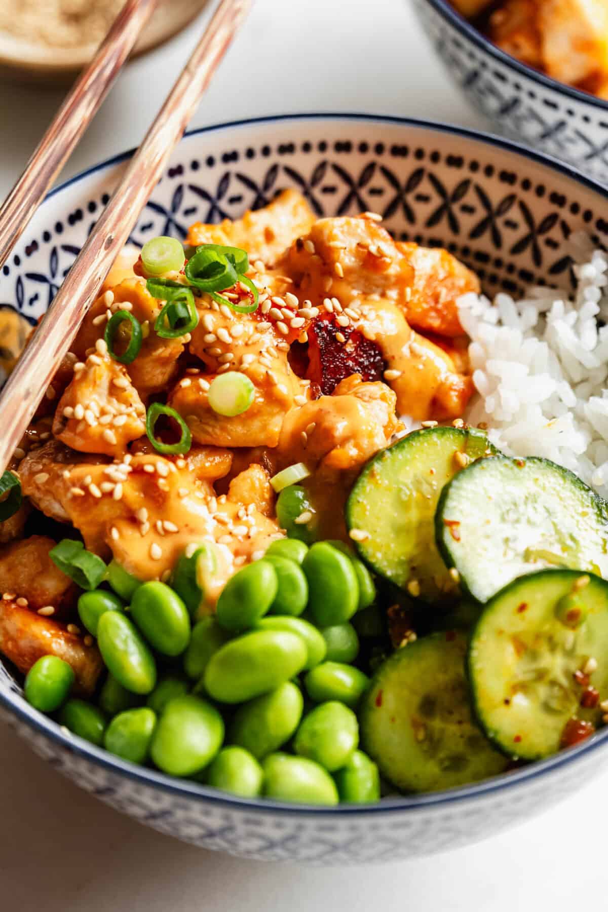 Close-up of a bang bang chicken bowl with rice, cucumber slices, edamame, and chopsticks resting on top.