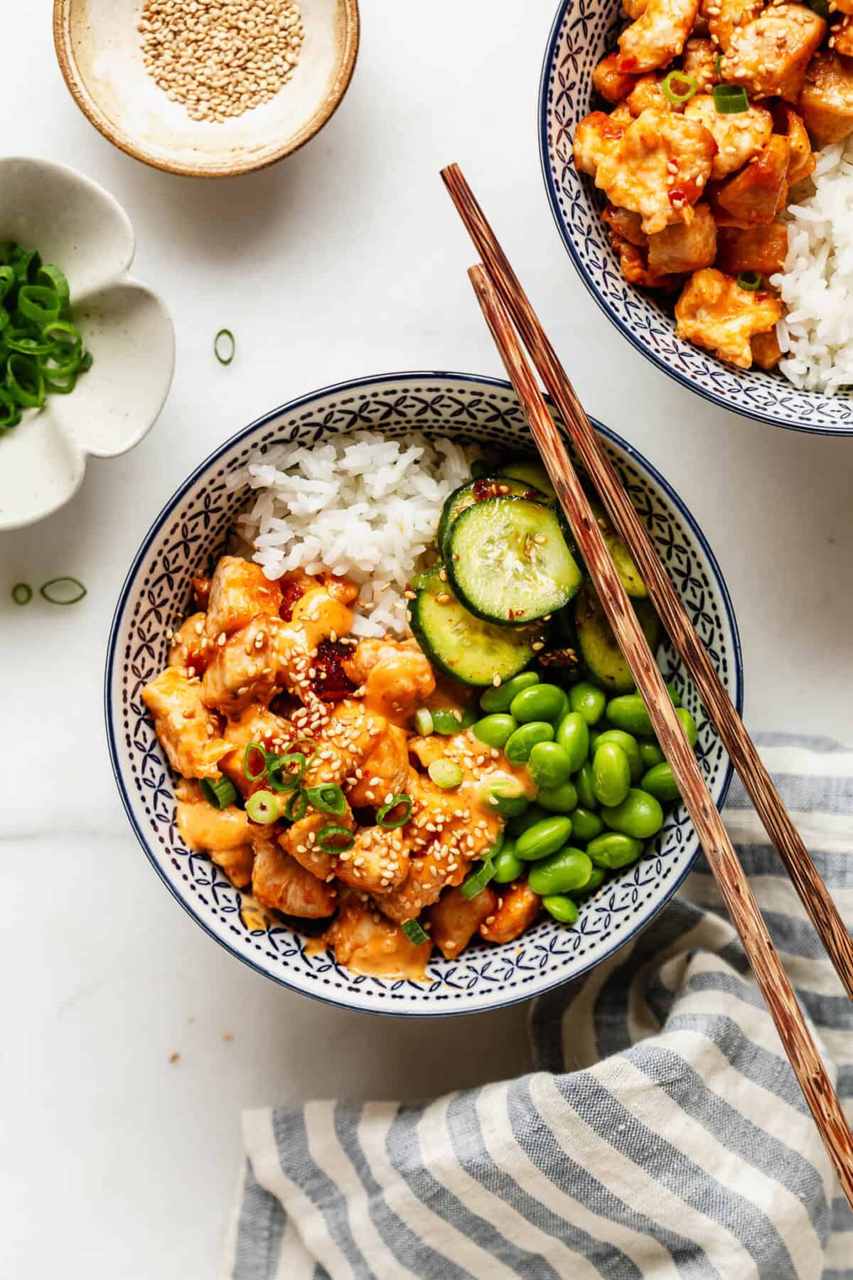 Two bang bang chicken bowls with rice and vegetables set on a table beside a striped linen napkin.