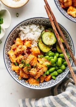 Overhead view of a bang bang chicken rice bowl garnished with sesame seeds and green onion, with a small dish of sesame seeds nearby.