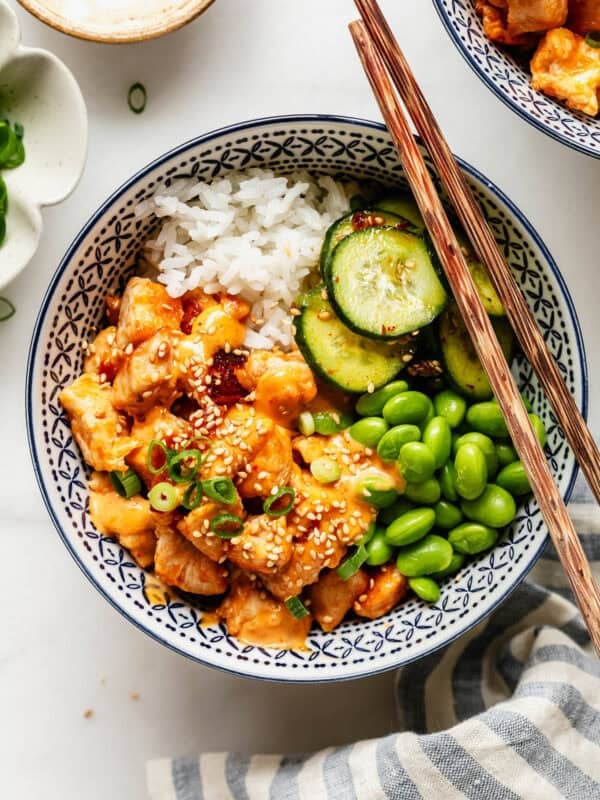Overhead view of a bang bang chicken rice bowl garnished with sesame seeds and green onion, with a small dish of sesame seeds nearby.