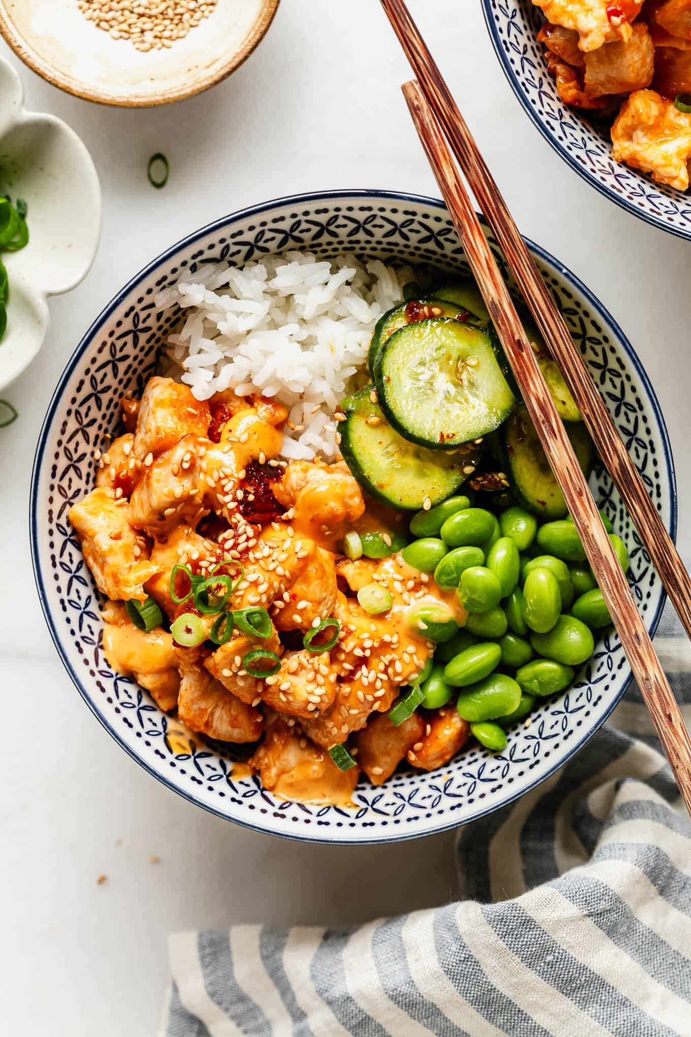 Overhead view of a bang bang chicken rice bowl garnished with sesame seeds and green onion, with a small dish of sesame seeds nearby.